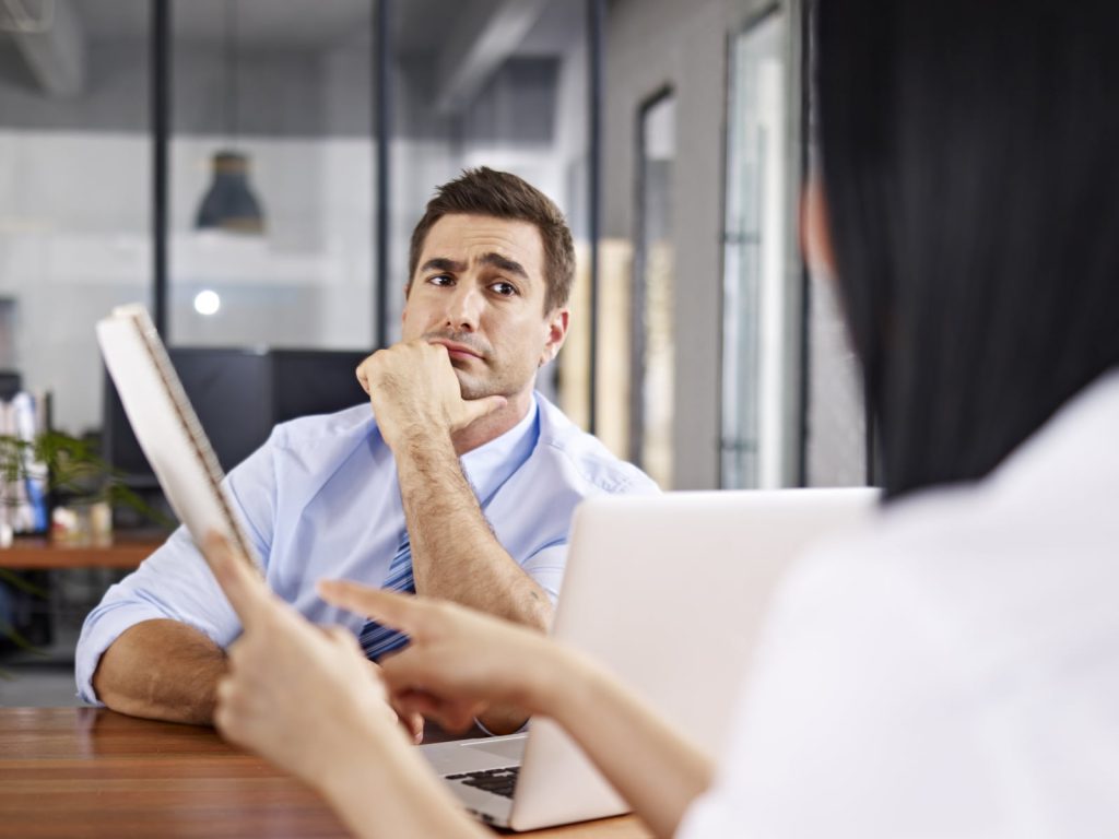 A man in a blue shirt sitting at a desk, looking thoughtful during a conversation, while a woman with a laptop gestures towards a document she is holding.