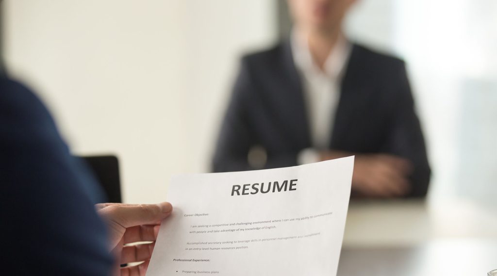 Close-up of a hand holding a resume during a job interview, with the resume in focus and the businessperson across the table slightly out of focus.