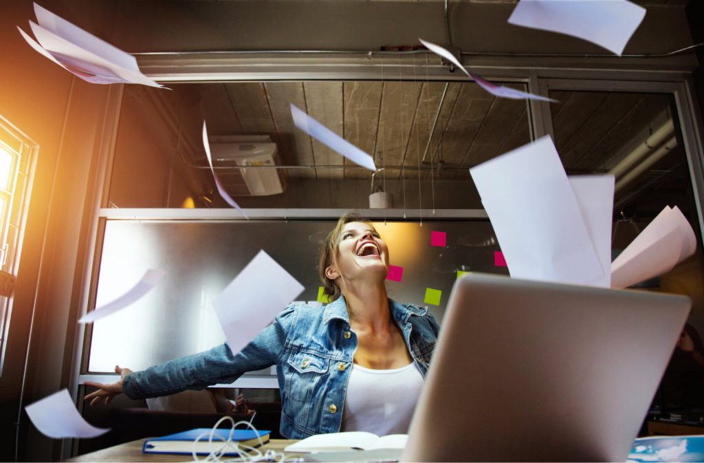 Businesswoman smiling at her desk as papers float around her, enjoying her work.