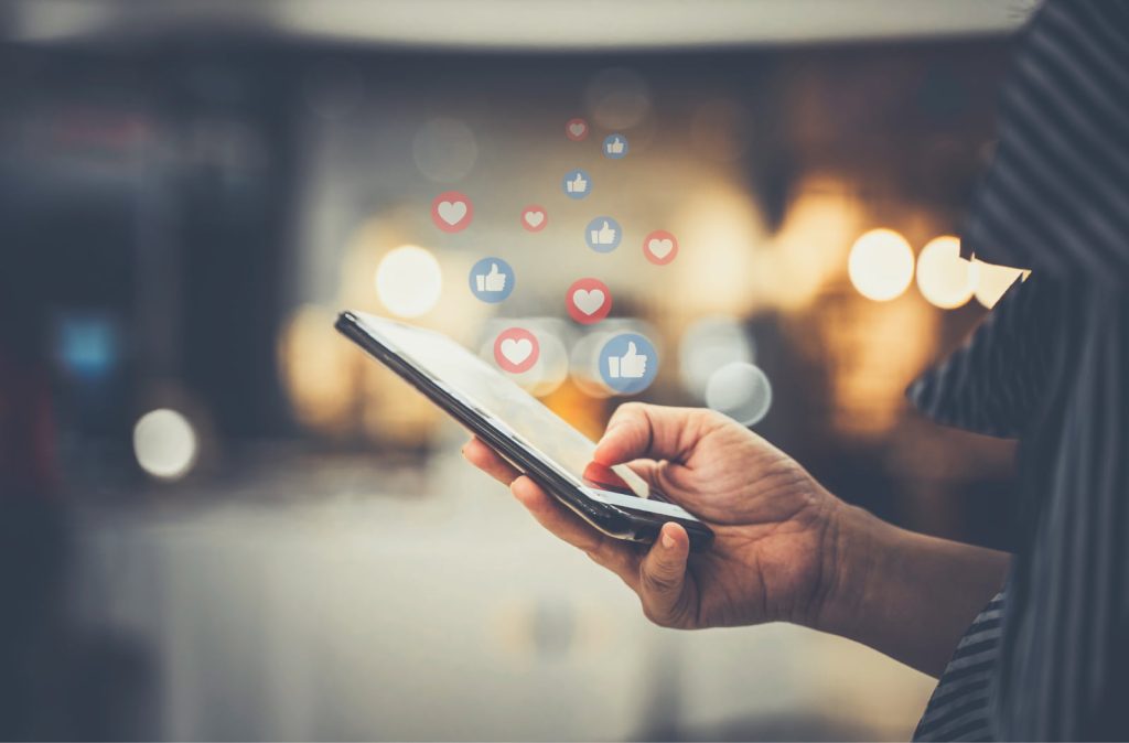 A hand holding a smartphone with floating heart and like reactions coming out of the screen, representing social media interactions.