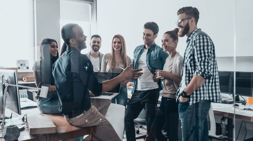 A diverse group of young professionals in an office, casually standing and chatting around a desk in a modern, open layout.