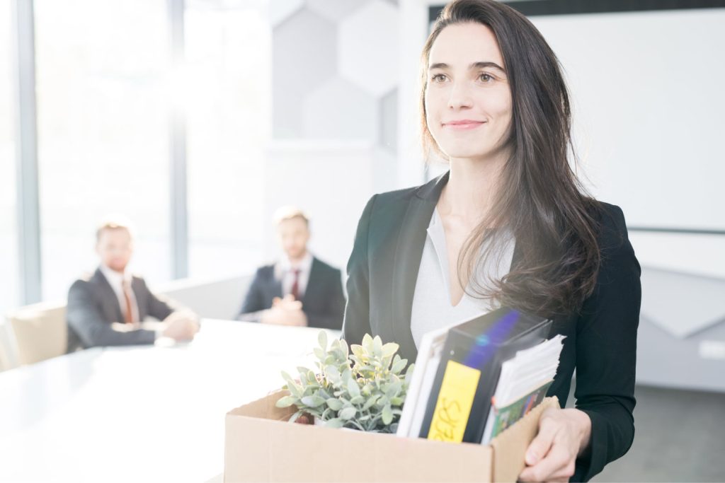 Smiling young businesswoman holding a box of personal belongings, leaving the office after quitting her job.