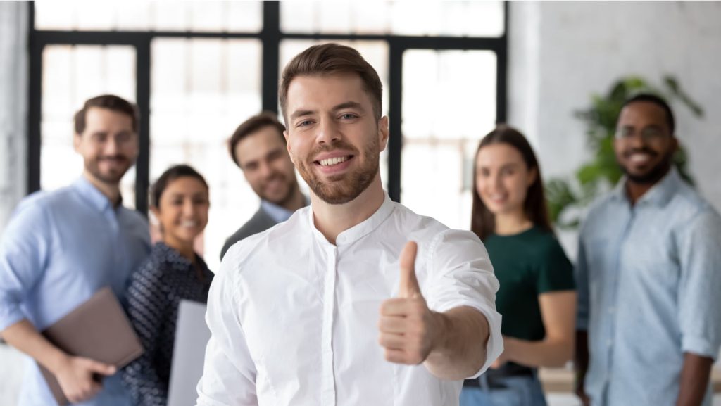 Smiling businessman giving a thumbs up with a diverse team of professionals standing in the background.