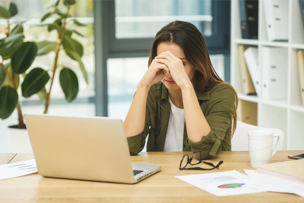 Frustrated young woman sitting at her desk, eyes closed, massaging her nose in a sign of stress and fatigue.