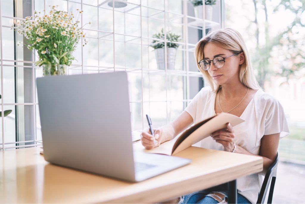 Female journalist in eyeglasses writing questions for an interview, using a textbook and attending an online webinar on her laptop.