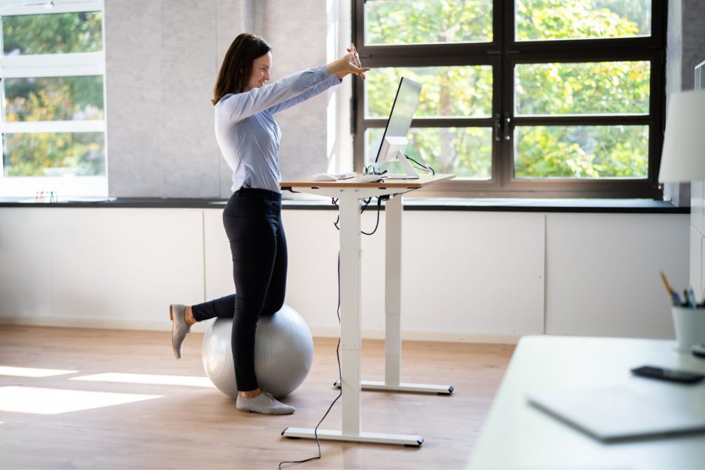 Woman performing a stretch exercise at a sit-stand desk, balancing one leg on an exercise ball in the office.