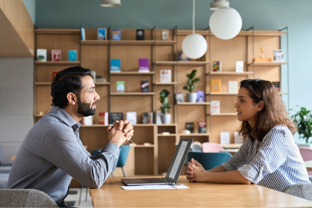 Indian male HR specialist listening to a female Latin candidate during a job interview in an office setting.