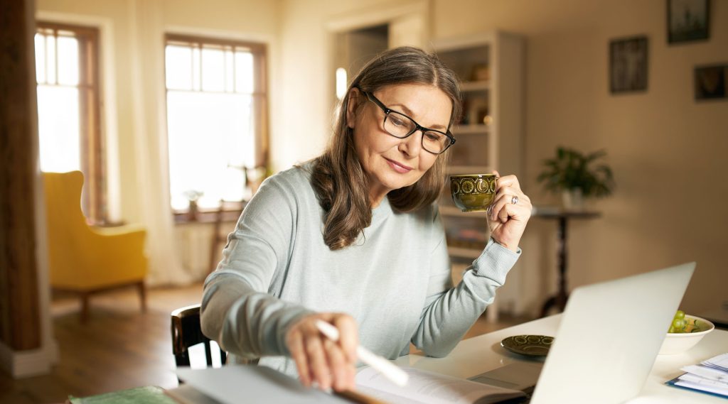 Serious, confident middle-aged female journalist sitting at a desk with a cup of coffee, books, and a laptop, making research and studying literature.