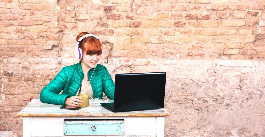 Young woman with headphones using a laptop at a home office, sitting at a small desk with a brick wall behind her, drinking a cold drink.