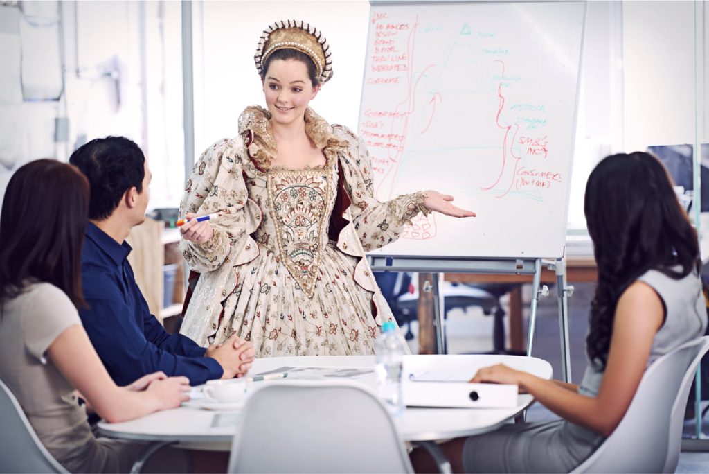 A woman dressed in a queen’s outfit giving a formal presentation in a modern office setting, creating a humorous contrast between her attire and the professional environment.