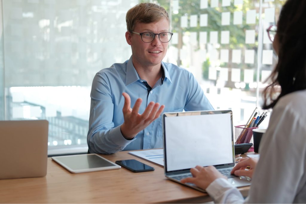 A man in a blue shirt sitting across from a woman with a laptop, engaged in a job interview discussion. The man gestures while speaking, and the woman is typing on her laptop.