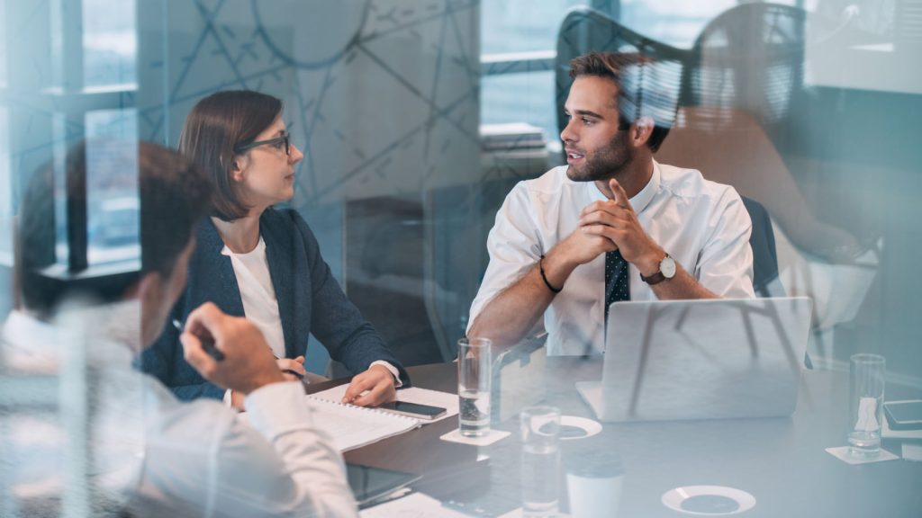 Group of business people talking around a table in an office boardroom, with a man and woman behind a glass wall.