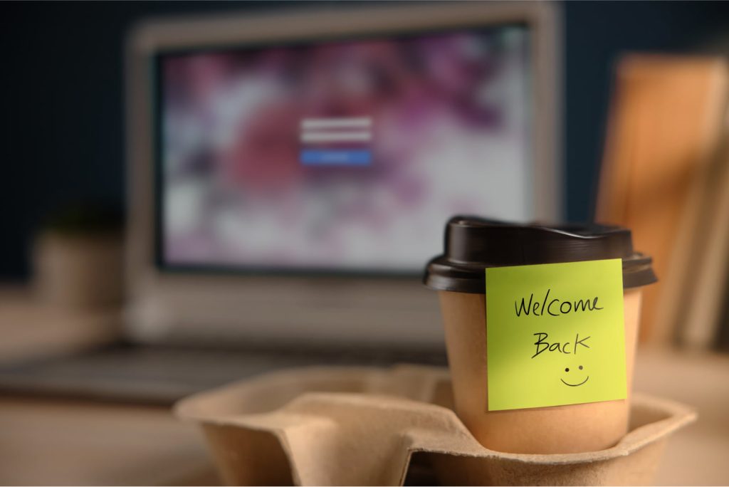 Closeup of a takeaway coffee cup with a note that says “Welcome back :)” on an office desk.