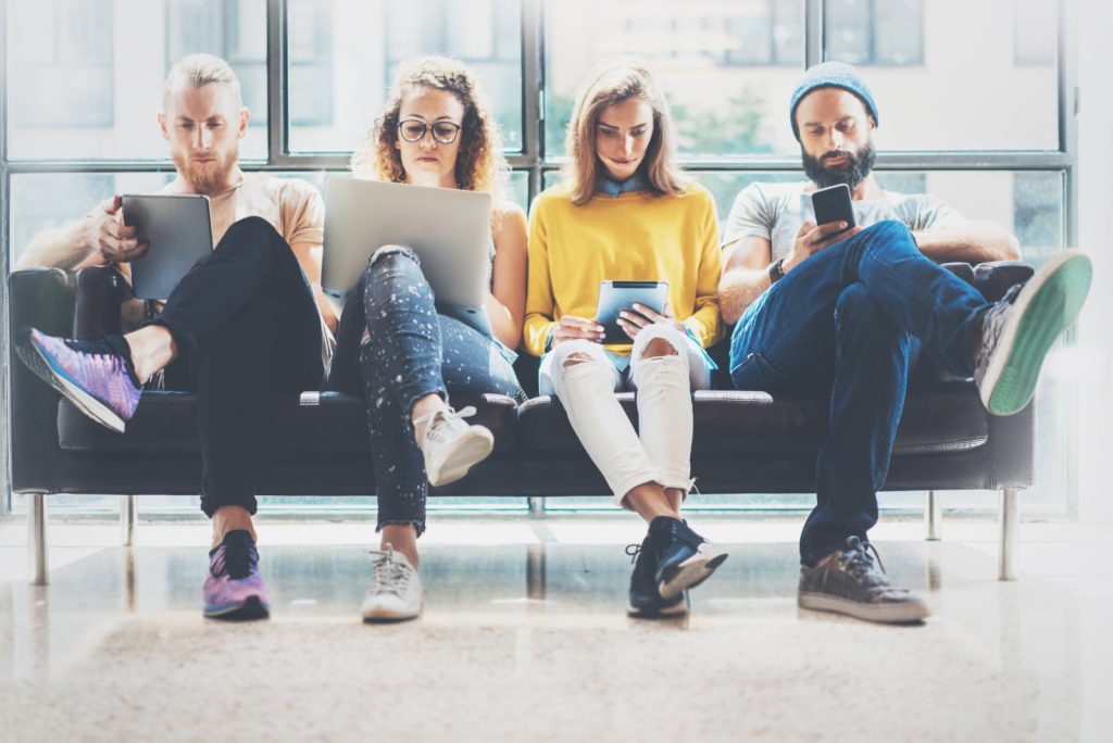 Group of adult hipsters sitting on a sofa, using modern gadgets while waiting.