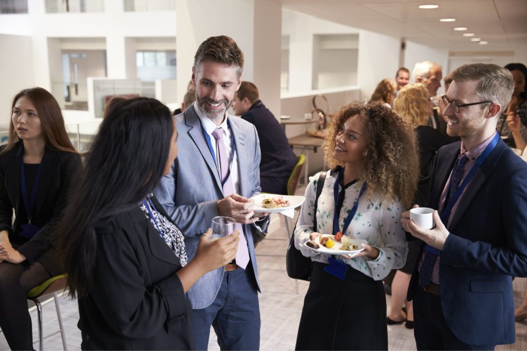 Group of delegates networking during a conference lunch break, standing with plates of food and drinks.