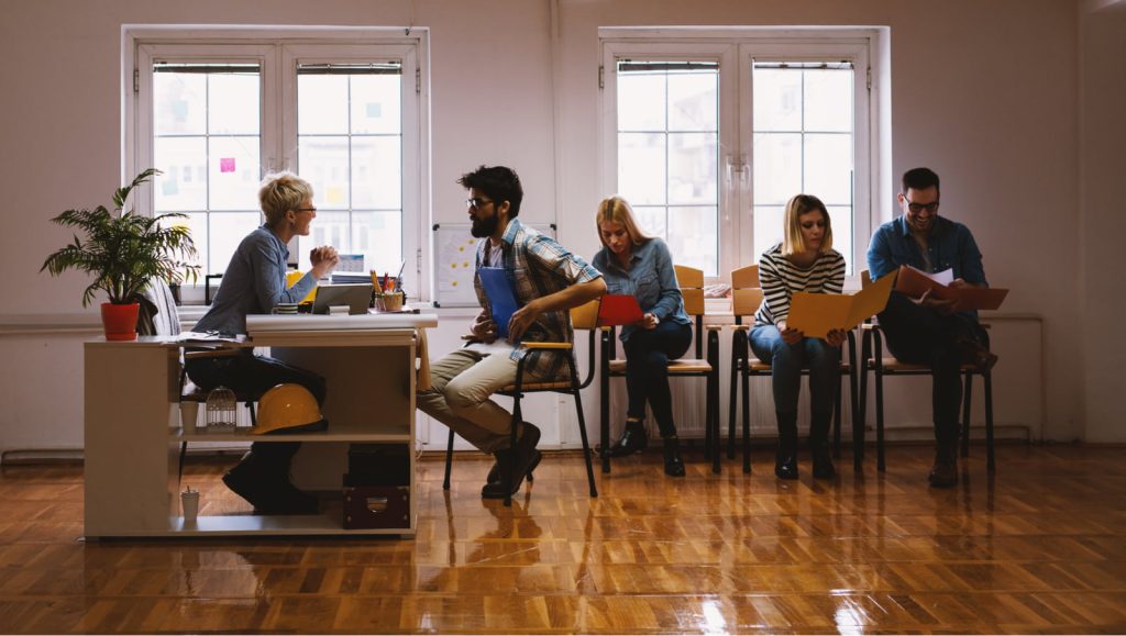 Four young people in a side view, seated in line with portfolios, while one is talking with an entrepreneur.