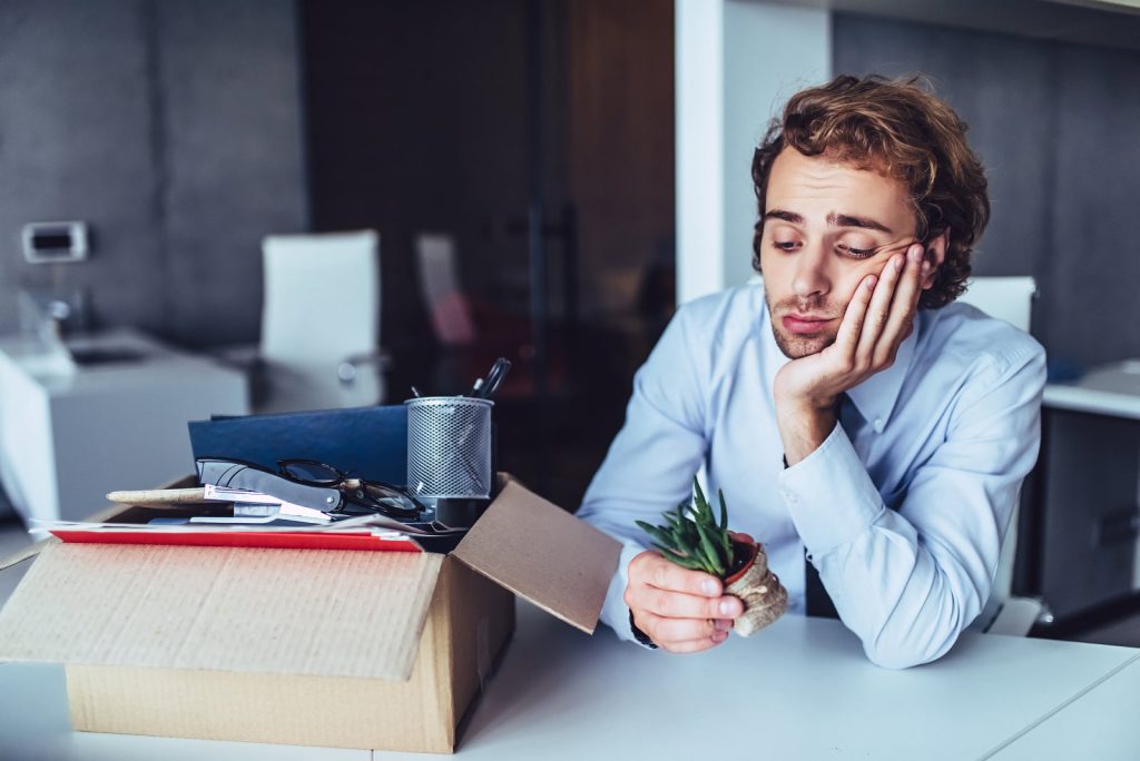 Man with a disappointed expression holding a plant next to a cardboard box of personal items after leaving his job.