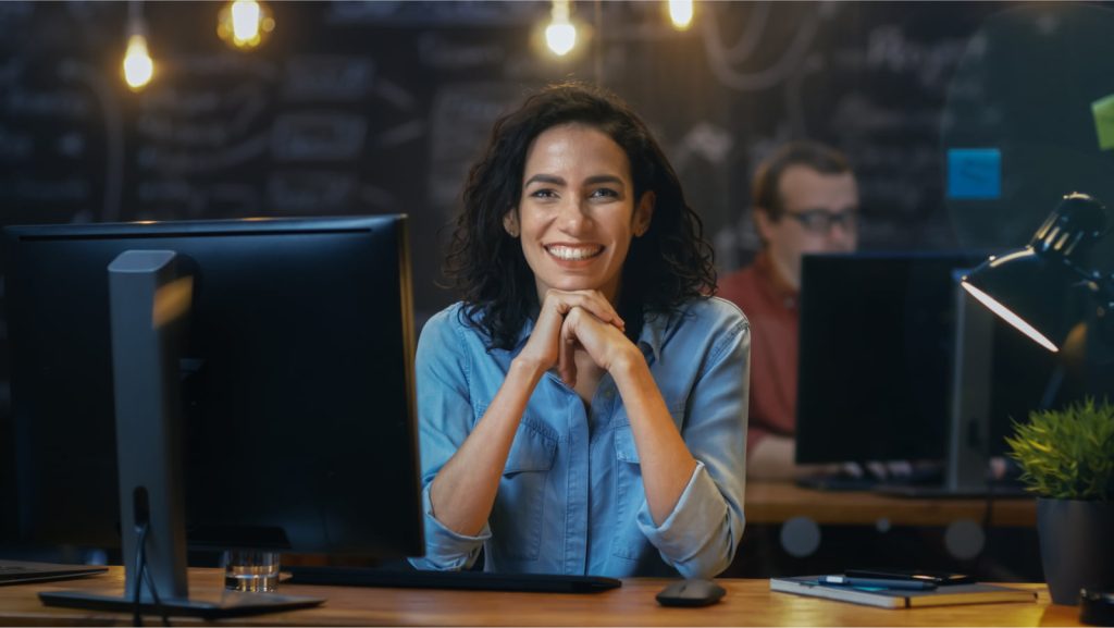 Smiling woman in a modern office sitting at her desk with hands clasped, giving off a positive and energetic vibe.