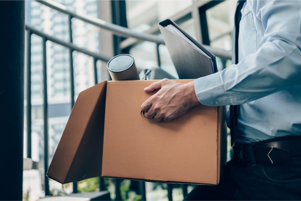 Man holding a cardboard box with a laptop bag, dossier, and drawing tube, symbolizing quitting or being fired from a job.