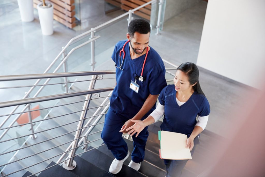 Two healthcare colleagues, a male and a female, wearing scrubs, talking on the stairs at a hospital. The woman is holding a folder.