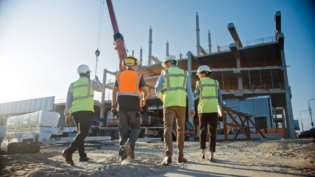 Diverse team of specialists, including a civil engineer, investor, and worker, inspect a commercial building construction site, with cranes and skyscraper formwork frames in the background.