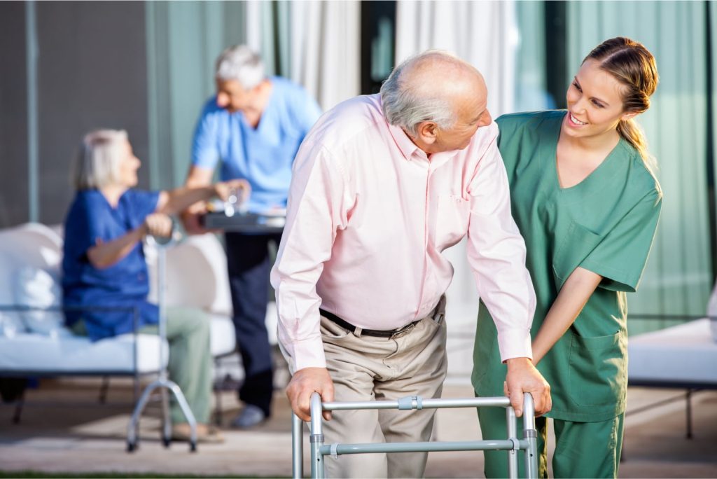 Happy female caretaker assisting a senior man with a Zimmer frame at a nursing home.