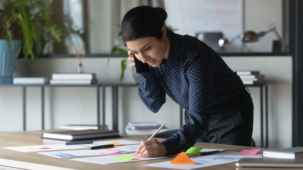 Serious Indian businesswoman talking on a cellphone while writing notes on colorful sticky papers in an office, working with financial documents and statistics.