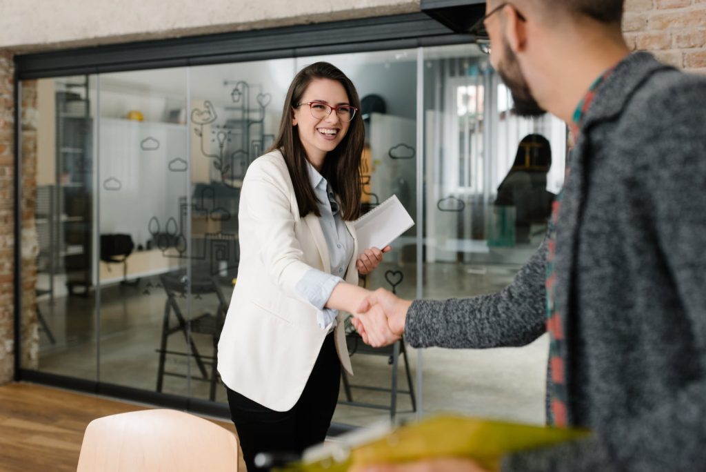 Two employers sitting in an office shaking hands with a young Asian woman after successful negotiations or interview, view from behind the employers.