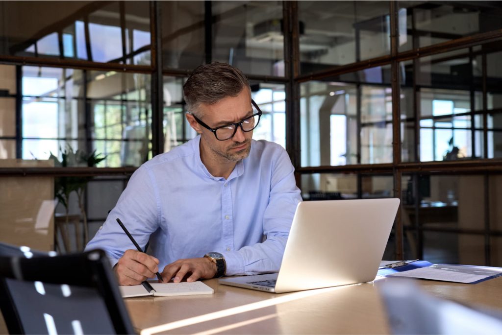 Mature businessman executive manager looking at a laptop, attending an online webinar or virtual meeting, and taking notes in an office.