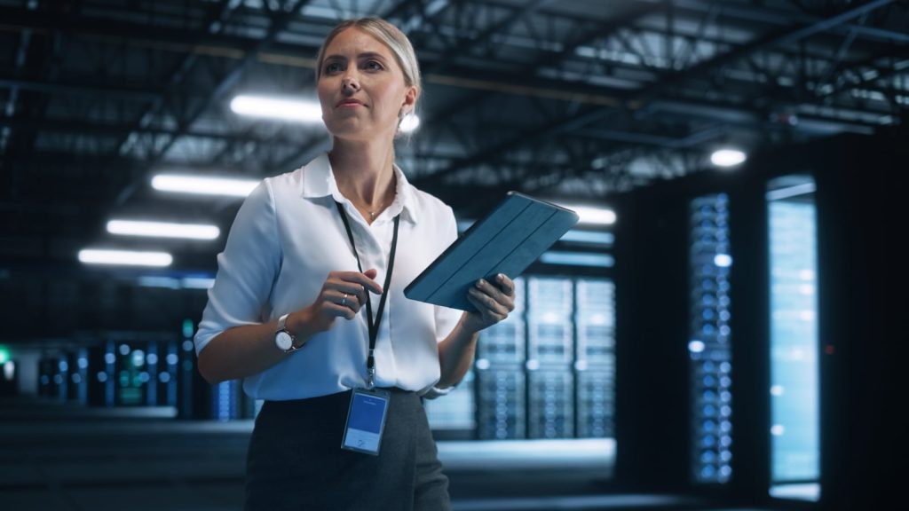 Female e-business entrepreneur using a tablet with a smile, wearing a white shirt and lanyard, in a data center with IT engineers.