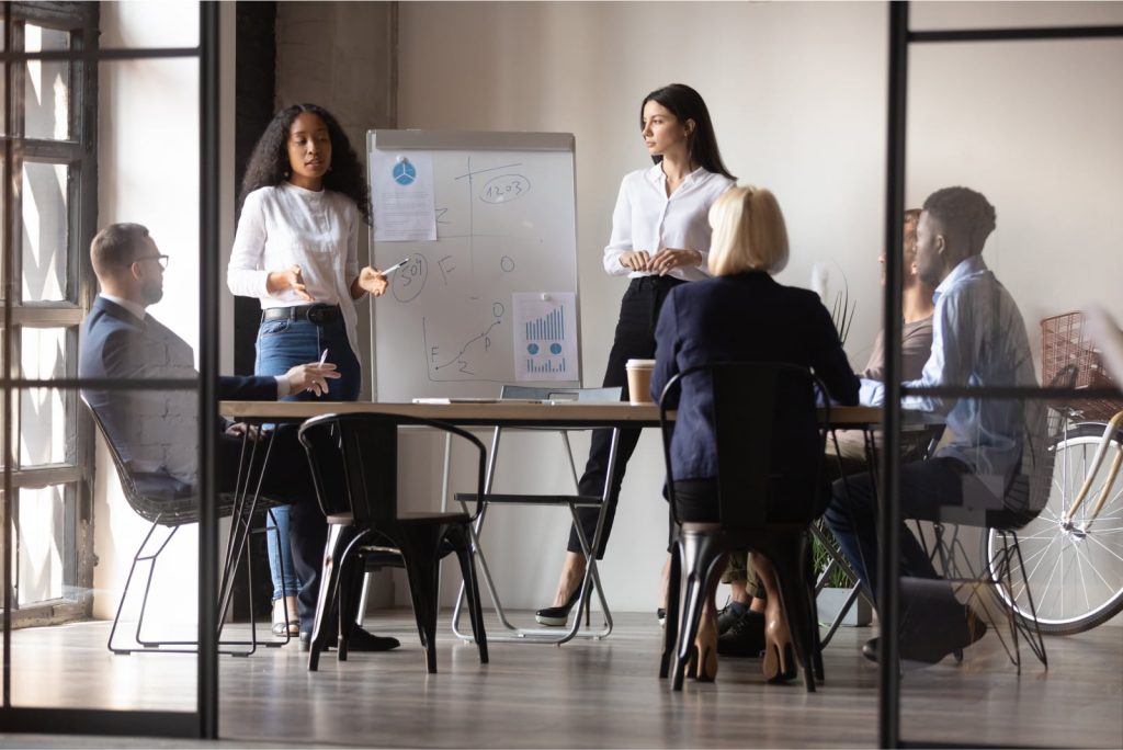 Diverse group of colleagues in a brainstorming session with two women presenting at a whiteboard.