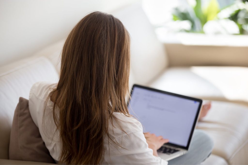 Woman sitting on a couch typing a cover letter on her laptop for a job application, view from over her shoulder.