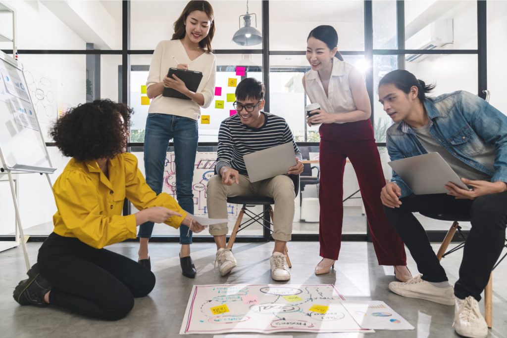 A group of five diverse young professionals collaborating in a creative planning session with notes and sketches on the floor.