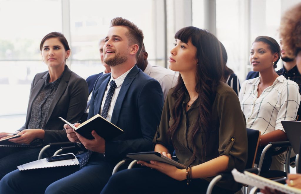 A group of professionals sitting and attentively listening during a workshop, with notebooks in hand, in a bright and modern setting.