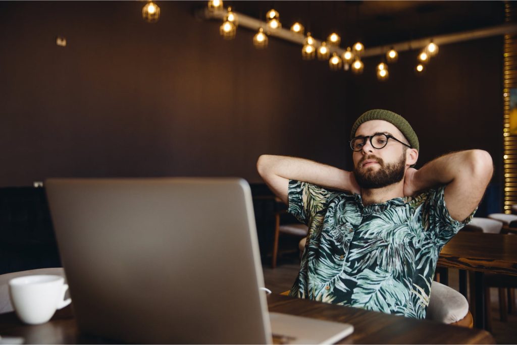 A man in a tropical shirt and beanie relaxes in a café with his laptop open in front of him, taking a break from work.