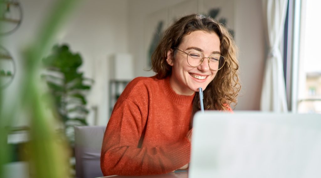 A young woman wearing glasses and an orange sweater is smiling while working on her laptop at home.