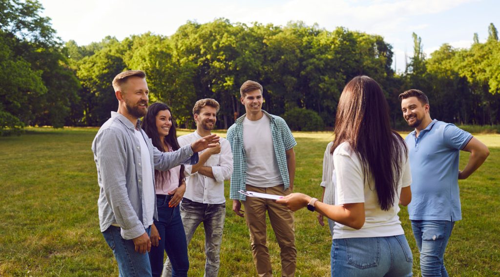 Group of happy friends or coworkers listening to a manager with a clipboard in a summer park, discussing outdoor team-building game tasks and rules.