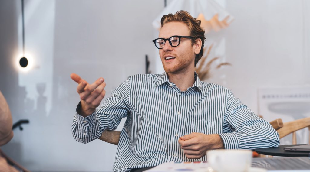 A man wearing glasses and a striped shirt gestures while talking in a casual office setting with a coffee cup on the table.