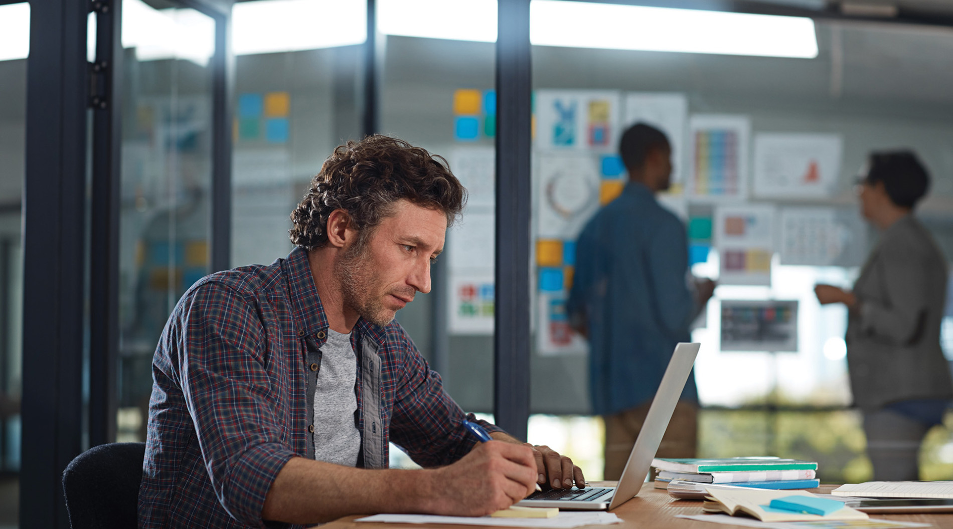 A man working on a laptop with notebooks and papers on a desk, while two colleagues stand and discuss work in the background in a modern office setting.