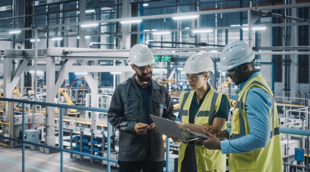 Three industrial workers wearing hard hats and safety vests stand together in a modern factory, looking at a laptop. The background shows a large manufacturing facility with machinery and equipment.