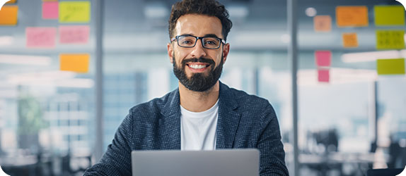 Smiling IT professional sitting at a desk with a laptop, representing innovation and technology-driven problem-solving in a collaborative office environment.