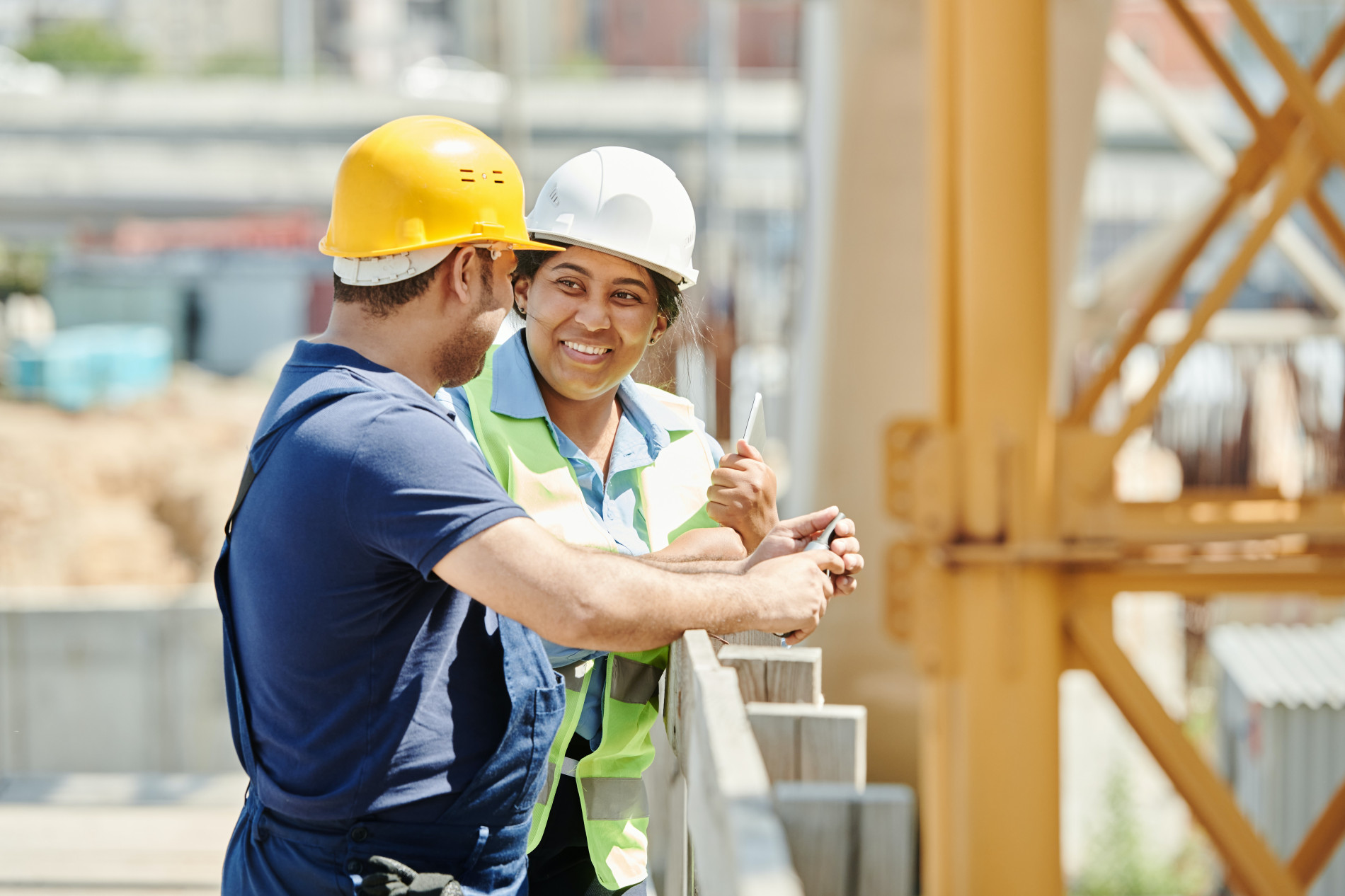 Two construction workers smile at one another on an indistinct work site.