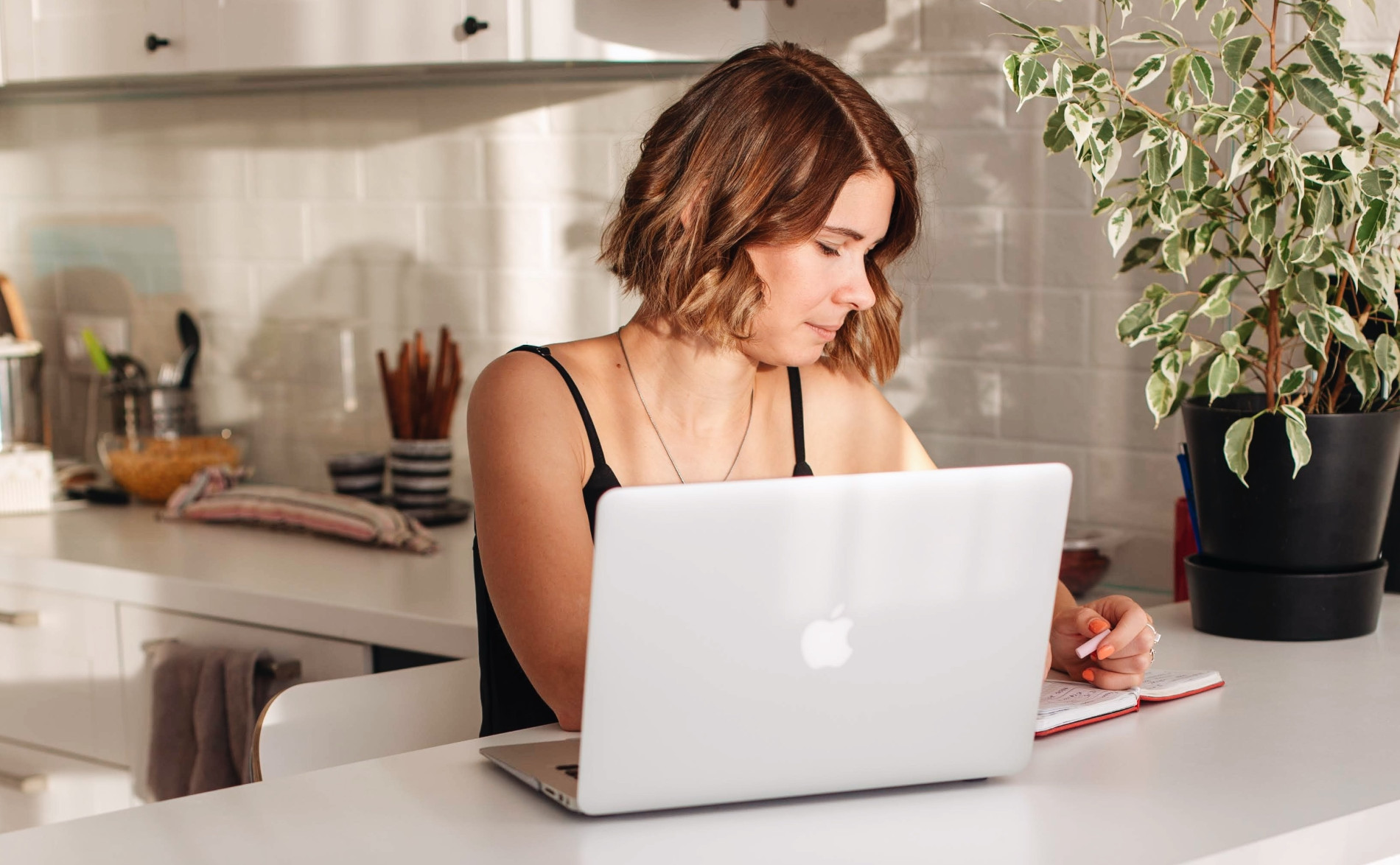 A female remote worker sits at a laptop in a bright, well-lit kitchen counter. 