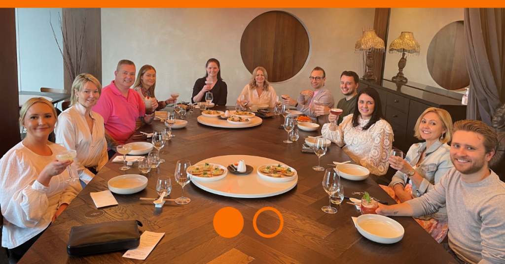 Group of individuals sitting around long wooden dining table with plates and glasses in front of them.