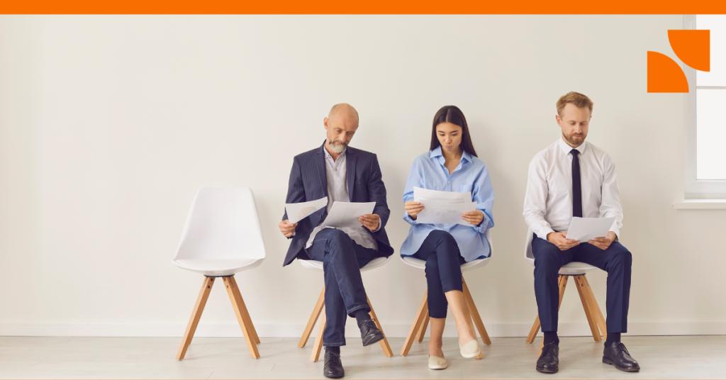 A row of chairs against a white wall with 3 individuals sitting in them reading papers.