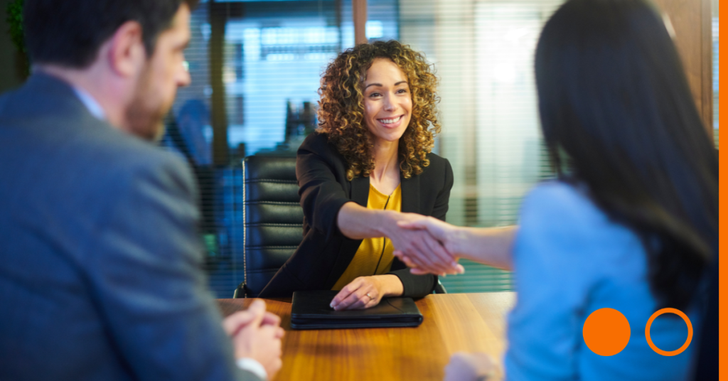 Woman in chair at desk shaking hands with an individual across the table with back to camera. There is another individual with back to camera as well.