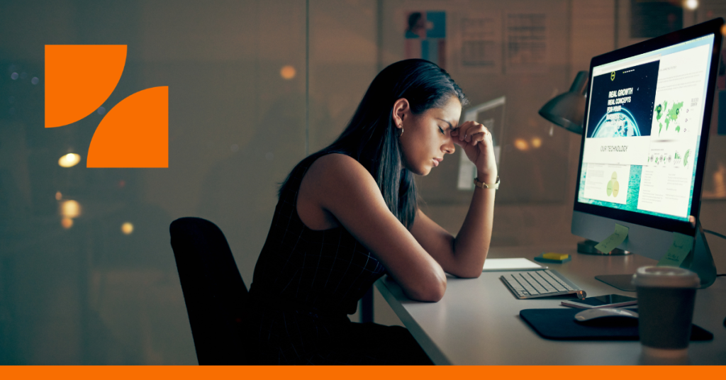 Woman at desk with large desktop computer display, hunched over with her eyes shut and pinching the bridge of her nose.