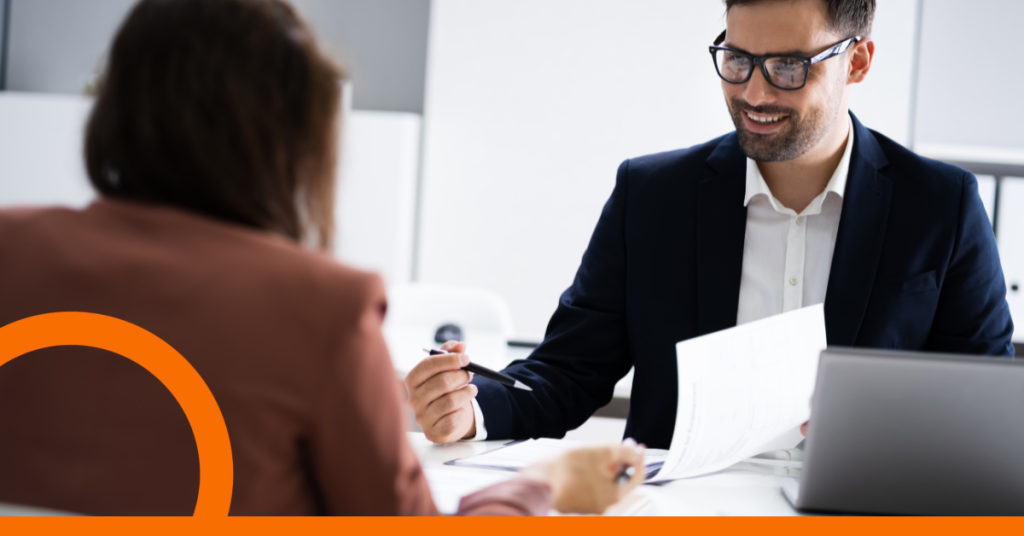 Man sitting at desk talking with individual with back turned to camera.
