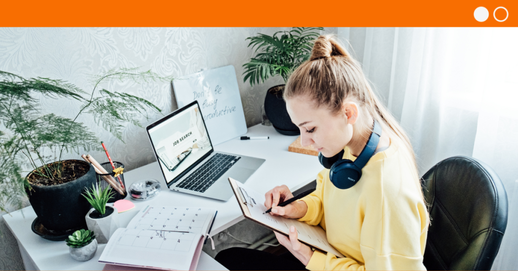 Woman with headphones around neck sitting at desk writing on a clipboard with laptop, diary, pens and whiteboard on desk in front of her.