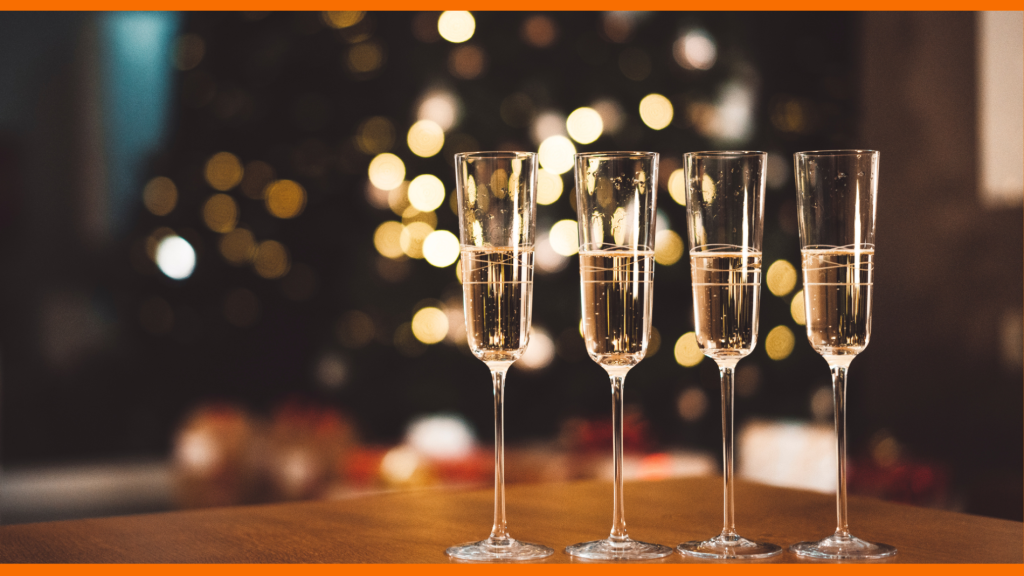 4 champagne flutes in a row on a table with a blurred christmas tree with lights in the background.